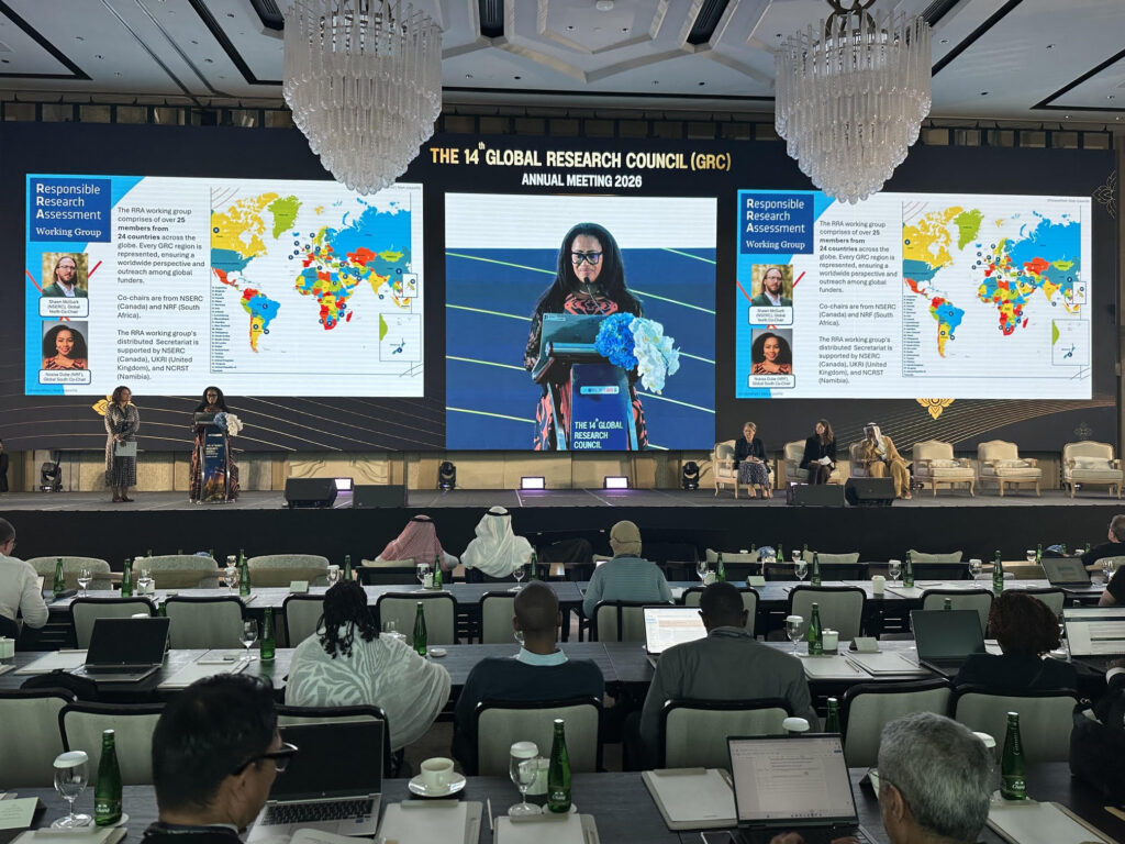 Wide shot of a conference hall during the 14th Global Research Council Annual Meeting 2026. Two speakers, Nosisa Dube and Giovanna Lima, stand on stage at a podium, while a large central screen displays a close-up of one speaker presenting. Slides on either side show information about the “Responsible Research Assessment Working Group” alongside a world map showing RRA WG members. Audience members sit at tables in the foreground with laptops and water bottles, facing the stage beneath large chandeliers.
