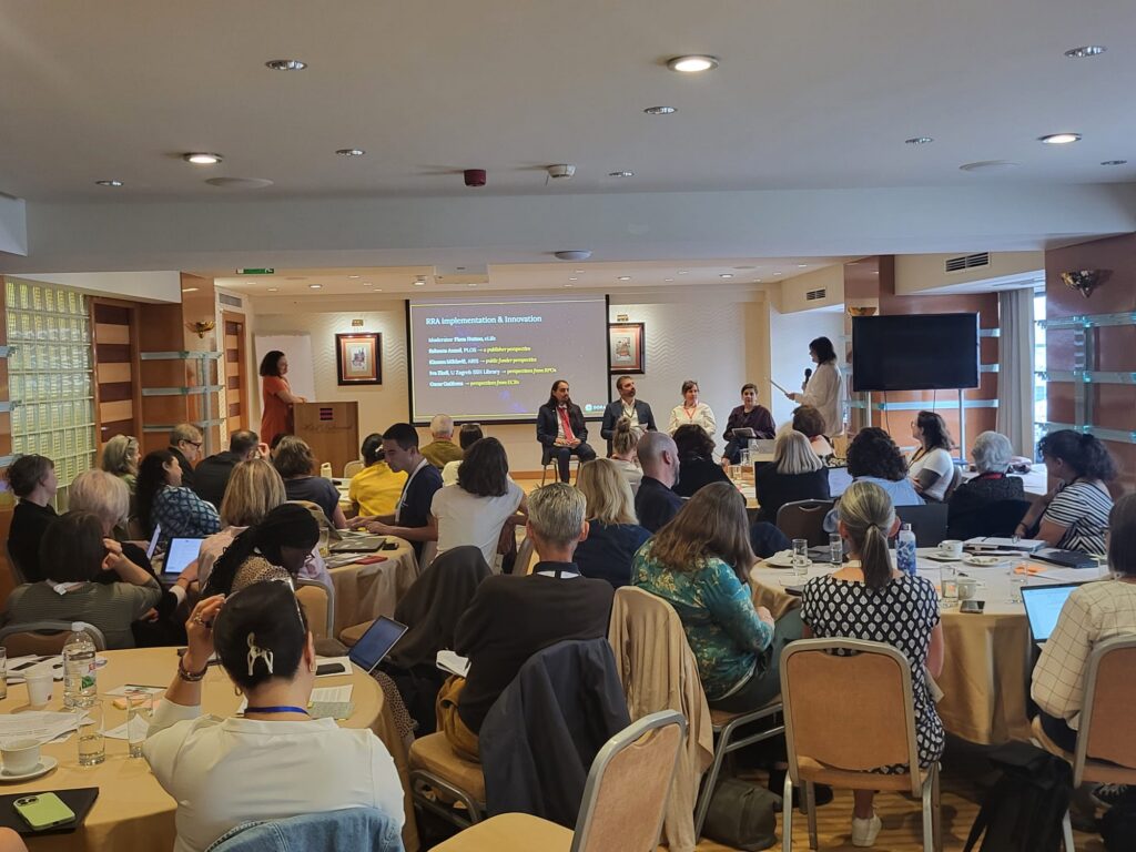 ZagrebWorkshop_Panel2 Workshop participants seated at round tables during a DORA event in Zagreb, with a panel of speakers discussing research assessment reform at the front of the room.